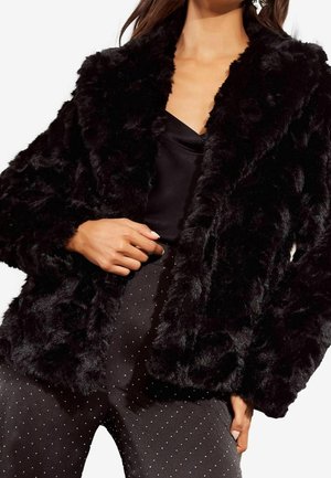 Femme portant une veste en fausse fourrure noire sur un haut en satin noir et un pantalon noir à petits pois blancs sur fond blanc.