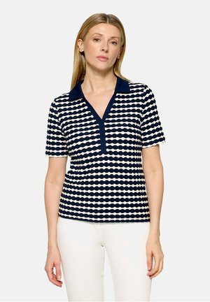 Femme portant un polo à manches courtes rayé bleu marine et blanc avec un col uni bleu marine et un pantalon blanc, debout devant un fond uni.