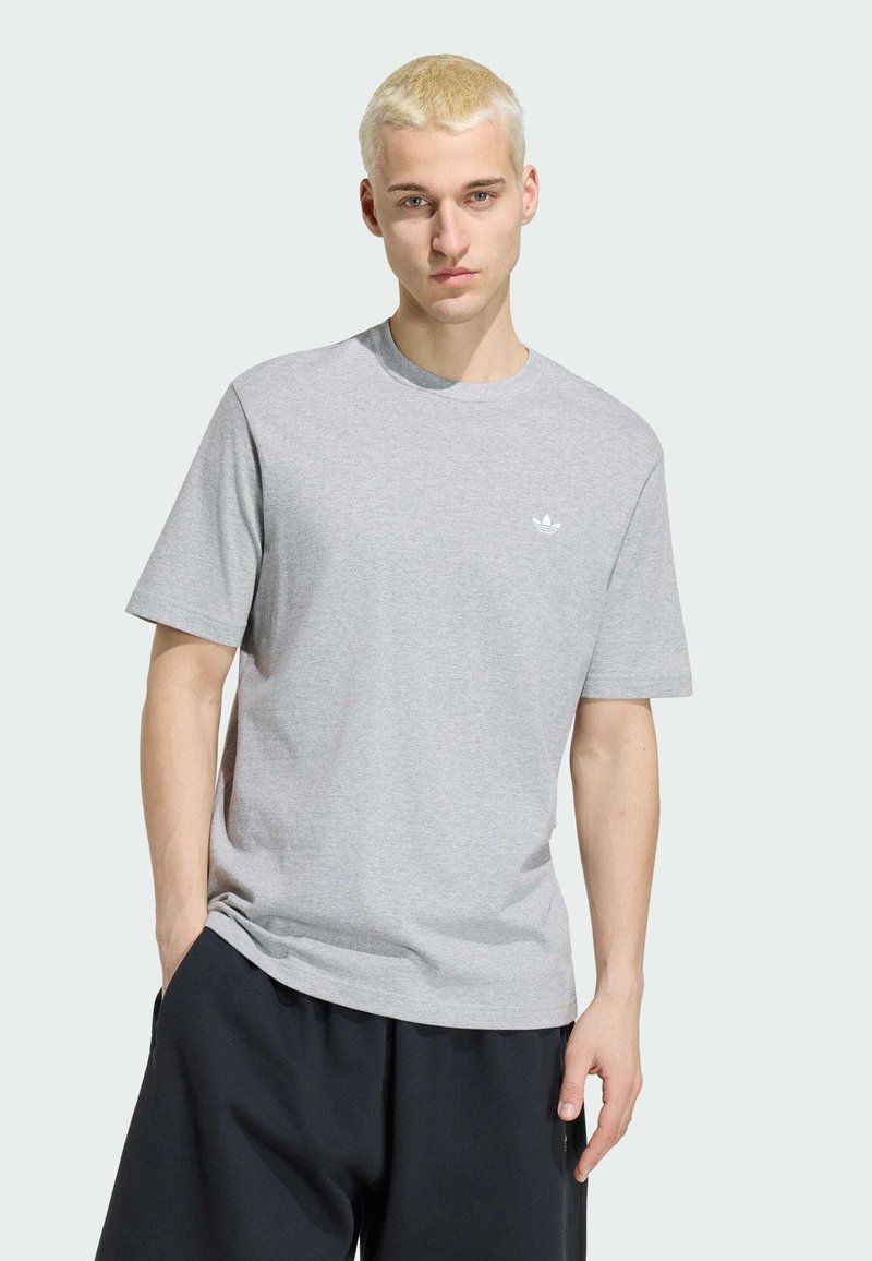Ung mand med kort blondt hår iført en lysegrå Adidas t-shirt og mørke shorts, står med den ene hånd i lommen.