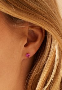 Boucles d'oreilles en forme de cœur en pierre précieuse rose serties en or. La pierre est taillée en facettes, capturant la lumière, complétant ainsi les cheveux lisses et de couleur claire.
