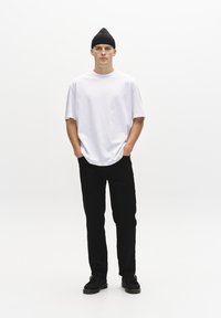 T-shirt oversized blanc, pantalon droit noir et bonnet noir. Le mannequin se tient sur un fond neutre, les mains dans les poches, avec des chaussures noires.