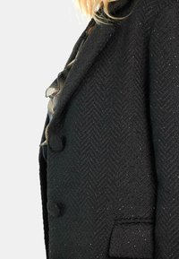 Abrigo negro en patrón de espiga con una tela texturizada. Presenta dos botones, un corte entallado y sutiles destellos a lo largo del material.