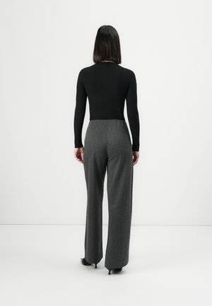 Kobieta z prostymi czarnymi włosami stoi tyłem, ubrana w czarną bluzkę z długim rękawem i szerokie szare spodnie na obcasach.