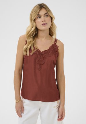 Femeie blondă purtând un top fără mâneci de culoare ruginie cu detalii din dantelă, asortat cu pantaloni albi cu accente brodate.