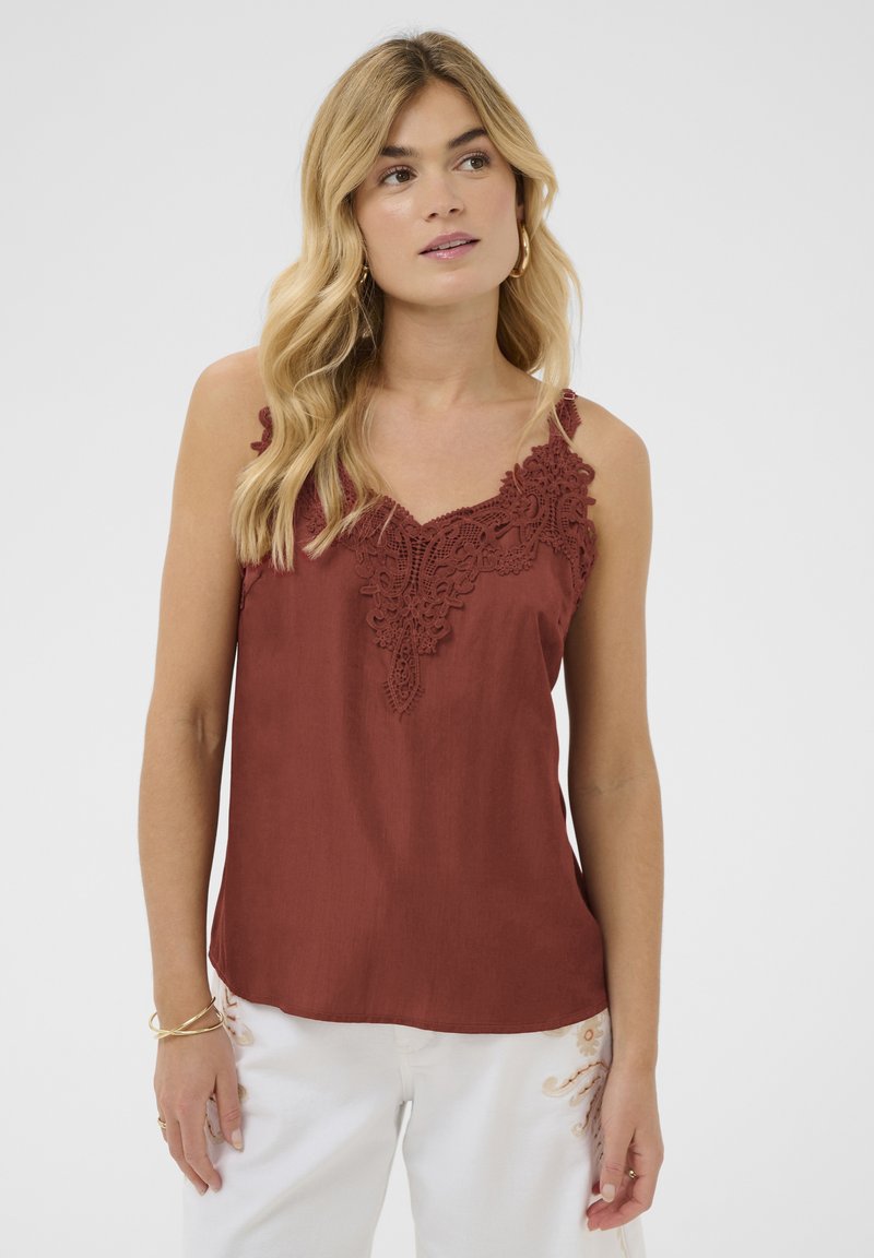 Femme blonde portant un haut sans manches de couleur rouille avec des détails en dentelle, associé à un pantalon blanc avec des accents brodés.