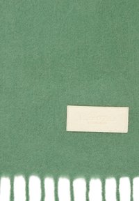 Couverture en laine verte avec une texture douce, dotée de franges blanches et d'une étiquette en tissu clair avec "Marc O'Polo" en relief.