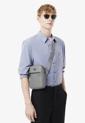 Bolso bandolera acolchado gris de diseño estructurado, que cuenta con un bolsillo con cremallera y una correa ajustable, llevado sobre una camisa azul claro.