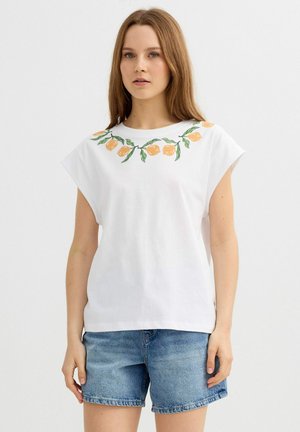 Joven mujer con camiseta blanca sin mangas con estampado de fruta de naranja y hoja verde, combinada con pantalones cortos vaqueros azules, de pie contra un fondo liso.