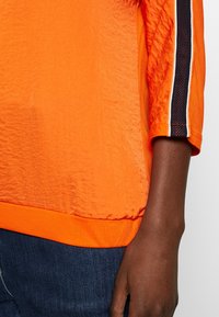 Oranger Pullover aus strukturiertem Stoff, mit Dreiviertelärmeln und kontrastierenden gestreiften Akzenten entlang der Ärmelenden.