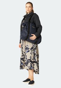 Granatowa torba puffer z pikowanym wykończeniem, z czarnymi paskami i akcentami z logo. Modelka nosi kwiecistą sukienkę i czarne buty typu slip-on.