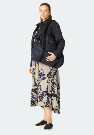 Marineblauwe gewatteerde tote bag met een quilted textuur, voorzien van zwarte straps en logo-accenten. Model draagt een bloemenjurk en zwarte instappers.