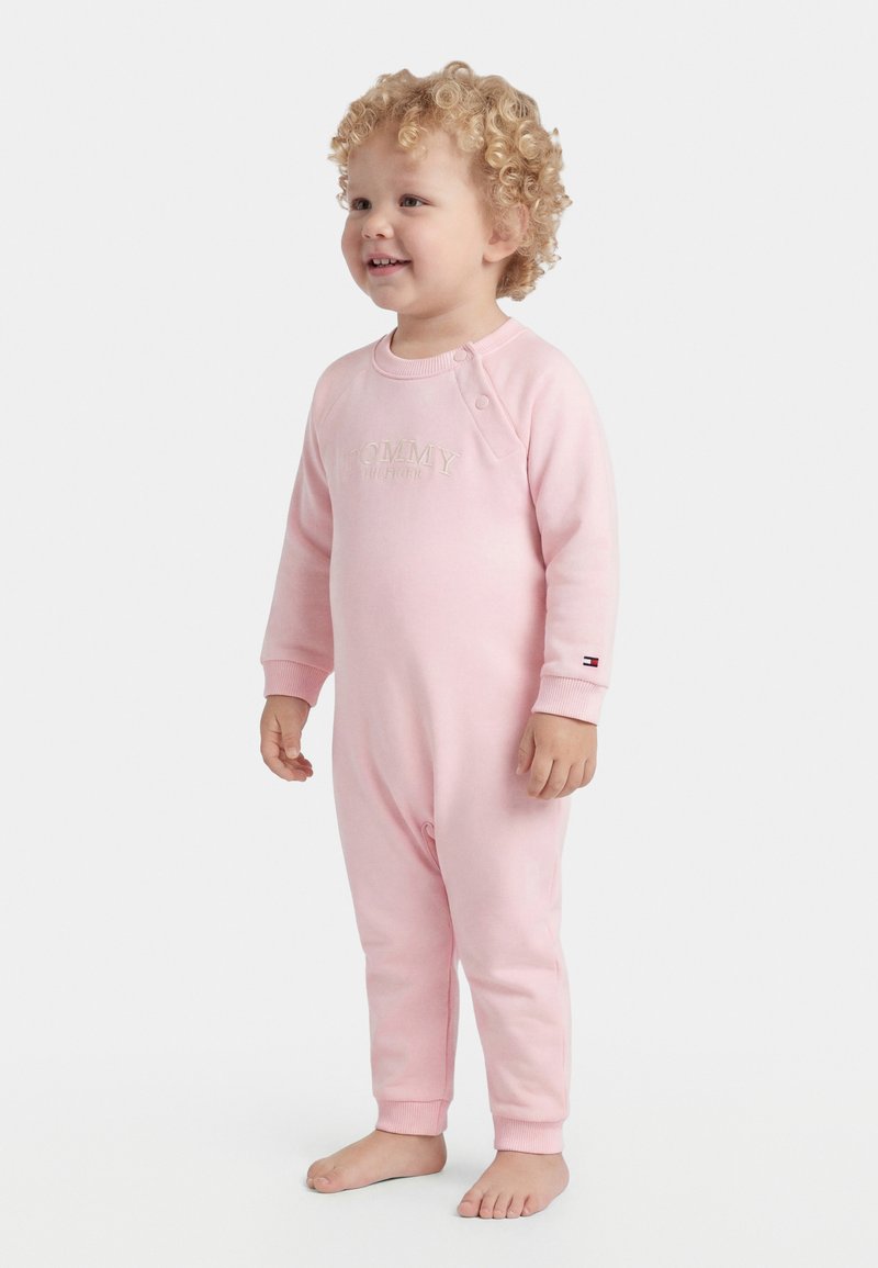 Νήπιο με σγουρά μαλλιά που χαμογελά, όρθιο ξυπόλητο, φορώντας ένα ανοιχτό ροζ ολόσωμο ρούχο με μακριά μανίκια και ριμπ μανσέτες.