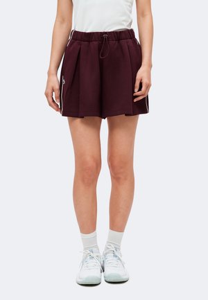 Personne portant un short plissé violet foncé avec un cordon, un haut blanc, des chaussettes blanches et des baskets blanches, debout devant un fond uni.