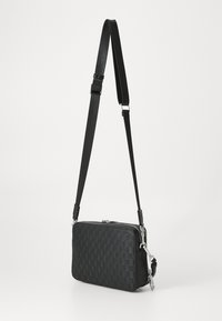 Sac bandoulière noir en matériau texturé, avec un motif géométrique, des fermetures éclair en argent et une sangle réglable.