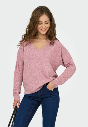 Jonge vrouw met krullend haar, gekleed in een roze V-hals trui en blauwe jeans, terwijl ze met één hand de handvatten van een zwarte tas vasthoudt en met de andere hand in haar zak zit.