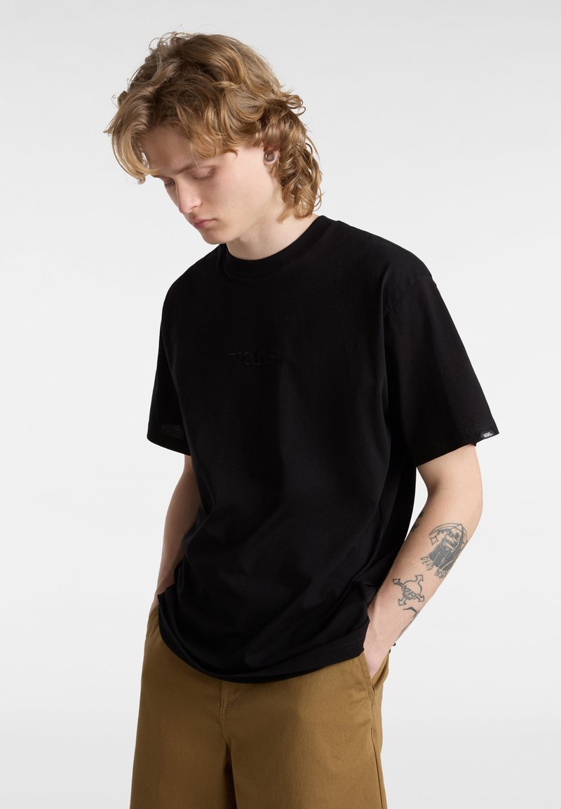 T-shirt in cotone nero con scollo rotondo, vestibilità rilassata e logo in rilievo sul petto. Indossato con pantaloni corti marroni, caratterizzati da una texture liscia.