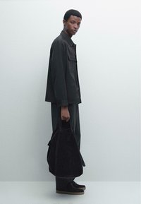 Giacca in tessuto scuro con due tasche sul petto, abbinata a pantaloni coordinati. Il modello tiene una borsa in suede scuro e indossa scarpe scure con suola chiara.