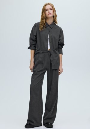 Chemise grise surdimensionnée avec col et boutons sur le devant, associée à un pantalon taille haute à jambes larges, tous deux en tissu texturé. Chaussures noires.
