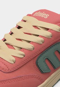 Κοντινό πλάνο σε κοραλί ροζ αθλητικό παπούτσι με μπεζ υφασμένους κορδόνια, τυρκουάζ ετικέτα με το λογότυπο "etnies" και το όνομα της μάρκας "etnies" στη γλώσσα.