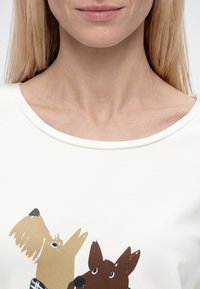 Gros plan d'une femme portant une chemise blanche avec un motif de dessin animé représentant deux chiens, un beige et un marron, sur le devant.