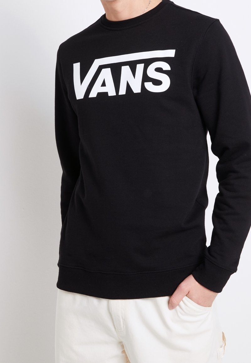 Persona che indossa una maglietta nera a maniche lunghe con un grande logo bianco "VANS" e pantaloni chiari, con una mano in tasca.