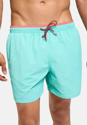 Hombre con pantalones cortos de baño turquesa con cintura coral y cordón negro y rojo, mostrado desde el torso hasta la mitad del muslo.