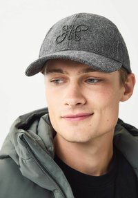 Grå tekstureret kasket med et logo, buet skygge og syede detaljer. Båret med en grøn pufferjakke, der har en lynlås ved halsen.