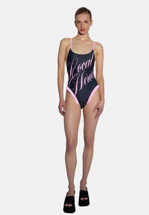 Kobieta w czarnym stroju kąpielowym z różowymi paskami i falbanami, z różowym napisem "Local Hero" na przodzie, oraz czarne sandały na platformie.