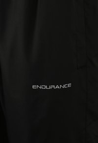 Svarta träningsbyxor i lätt material har en slät yta och en vit logotyp som säger "ENDURANCE" på sidan.