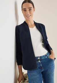 Námořnická bombardovací bunda z texturované látky se zapínáním na zip, doplněná bílým tričkem s výstřihem do V a modrými džínami. Modelka stojí u zdi.