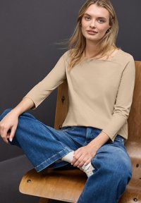 Top beige a maniche lunghe con scollatura ampia abbinato a jeans a gamba larga blu. Il modello è seduto su una sedia di legno, mostrando una posa rilassata.