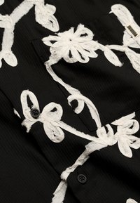 Tissu noir avec broderie florale blanche texturée, montrant un gros plan sur des boutons et une poche avec une petite étiquette en tissu.