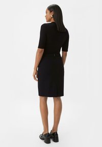 Robe noire ajustée à manches courtes, avec une fente dans le dos, texture lisse et détails de coutures subtils. Assortie à des chaussures à talons noires.