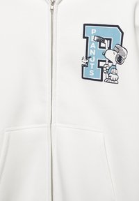 Witte hoodie met ritssluiting met een blauw-zwart beeld van Snoopy met een hockeystick naast de tekst "PEANUTS." Gemaakt van zacht katoen.