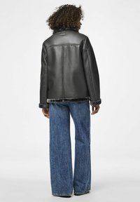 Giubbotto in finta pelle nera con colletto foderato in morbido pelliccia, vestibilità rilassata e dettagli di cucitura visibili. Abbinato a jeans in denim blu a gamba larga.