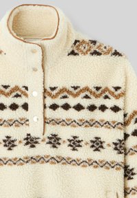 Pull à col haut en polaire crème, orné de motifs géométriques marron, avec trois boutons en métal argenté à la nuque et une finition texturée.