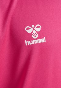 Rosa Sporttrikot mit glatter Textur, das ein weißes Hummel-Logo und ein Bienenmuster zeigt, zentral auf dem Stoff platziert.