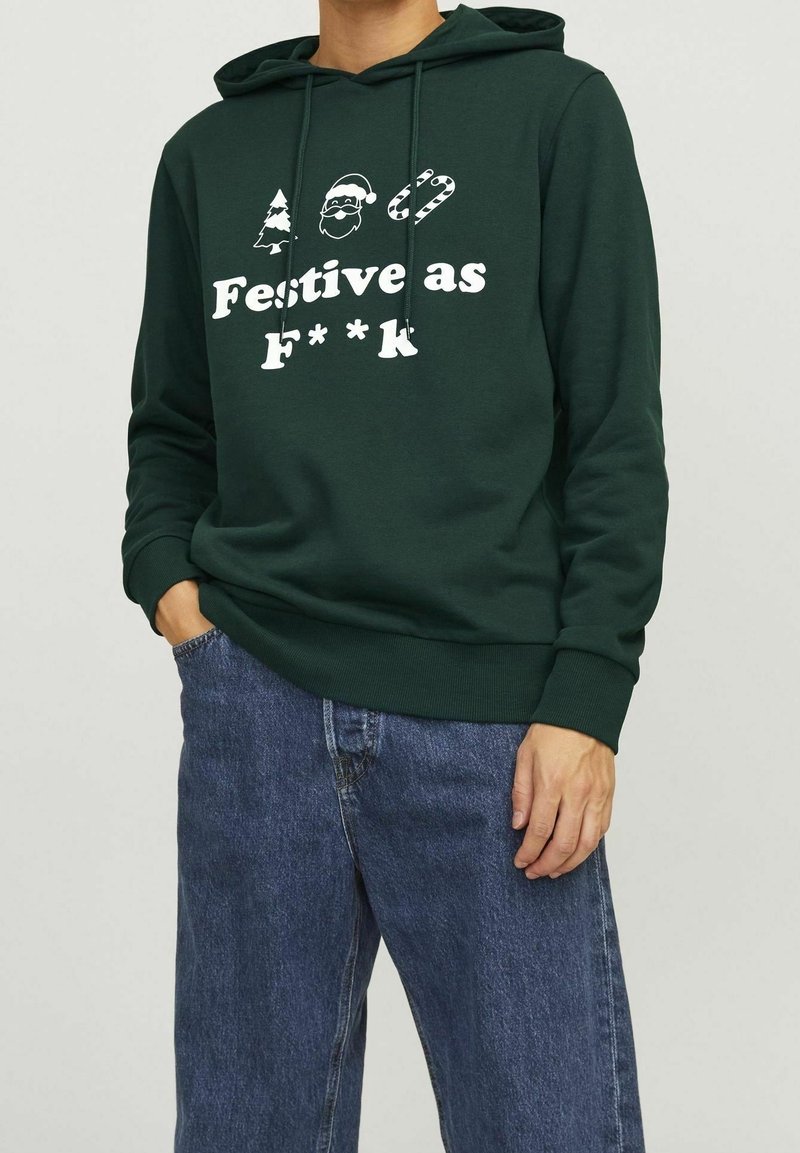 Personne portant un sweat à capuche vert foncé avec des icônes festives et le texte "Festif comme c**," assorti à un jean bleu.