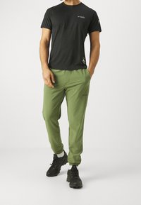 Man som bär en svart Columbia-t-shirt, olivgröna Columbia-joggingbyxor, vita strumpor och svarta terränglöparskor, stående med en hand i fickan.