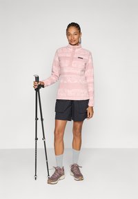 Lys rosa fleece-pullover med geometriske mønstre, halv-zip krage, svarte detaljer. Sammen med mørke shorts, grå sokker og tursko.
