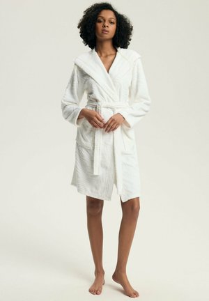 White robe made of textured fabric with a shawl collar, belt, and subtle leaf pattern. Knee-length with long sleeves and side pockets.
