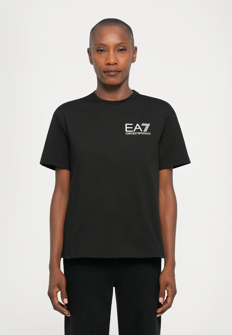 Person som har på seg en svart kortermet EA7 Emporio Armani t-skjorte, som står mot en ensfarget lys bakgrunn.