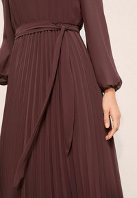 Bordeauxfarbenes, plissiertes Kleid mit langen Ärmeln und einer Taillenbindung. Der Stoff wirkt leicht und strukturiert mit vertikalen Falten.