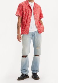 Camisa roja de manga corta con patrón floral blanco, combinada con jeans rasgados de color azul claro y zapatos negros con cordones.