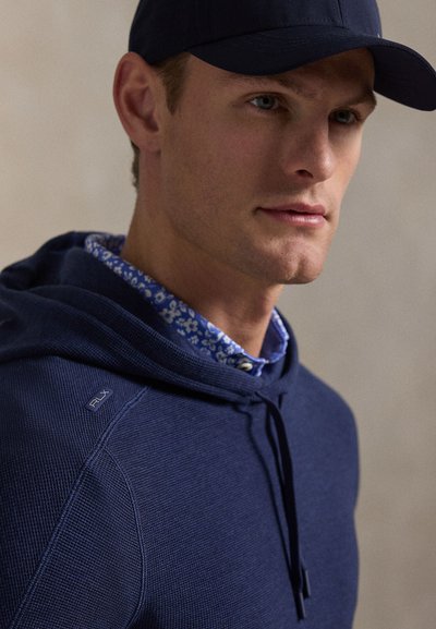 Ναυτικό μπλε υφασμάτινο φούτερ με κορδόνι και λογότυπο PLX, φορεμένο πάνω από μπλε πουκάμισο με λουλούδια και γιακά. Ρυθμιζόμενο κουκούλι και άνετη εφαρμογή.