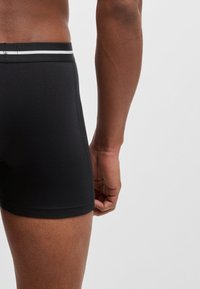 Black boxer shorts with a fitted design and elastic waistband featuring a thin white stripe. Smooth fabric with a soft texture.