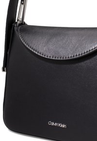 Borsa a tracolla in pelle nera con pattina arrotondata, hardware argentato e logo in rilievo. Superficie testurizzata con cuciture visibili.