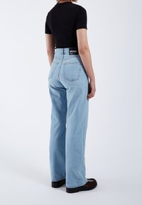 Personne portant un jean taille haute bleu clair, coupe droite, un haut noir à manches courtes et des chaussures noires, debout de dos sur fond blanc.
