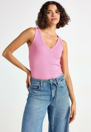 Kvinna med lockigt hår som bär en rosa ärmlös topp och högmidjade blå jeans med ett knytet bälte, stående mot en enhetlig bakgrund.