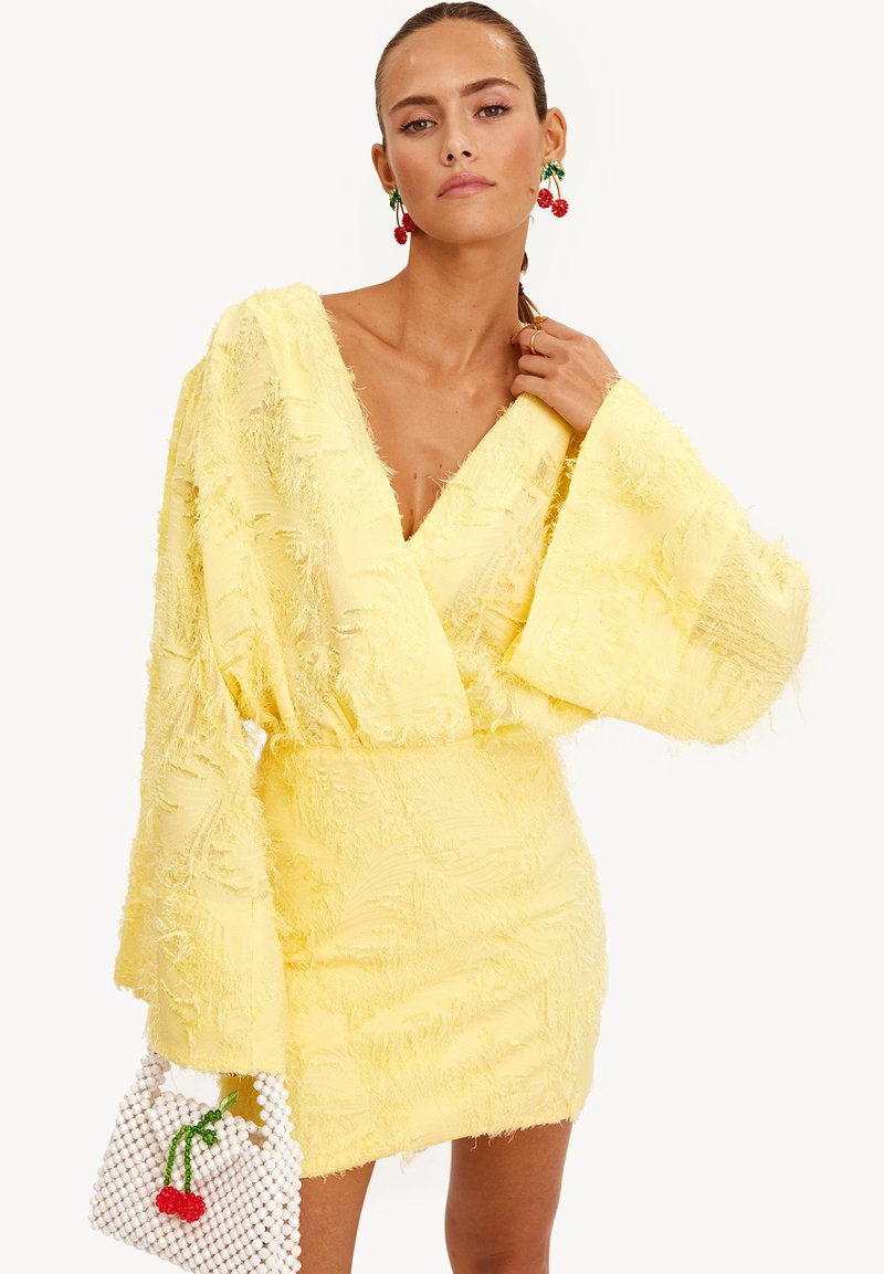 Un vestido mini amarillo con un acabado texturizado, escote en V y mangas largas acampanadas. La modelo sostiene un bolso blanco de cuentas con un diseño de cereza.